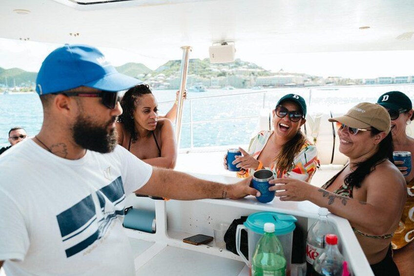 Un grupo de personas en un bote, sosteniendo bebidas, con una masa de agua y colinas al fondo. 4