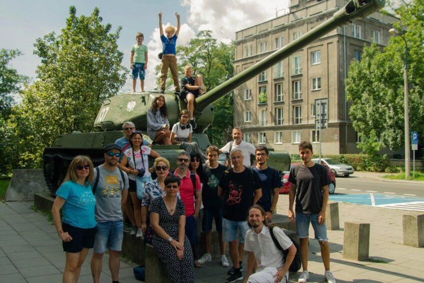 A group of people posing in front of a military tank, with a building and trees in the background. 3