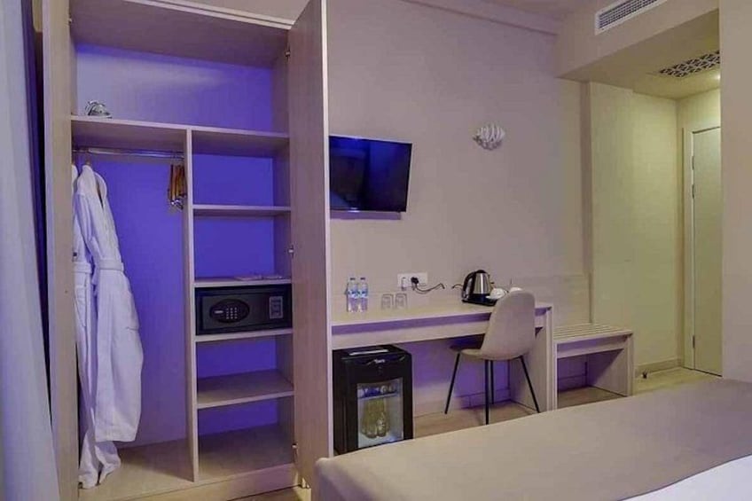 A hotel room with a bed, a desk with a chair, a television, a minibar, and a wardrobe. 3