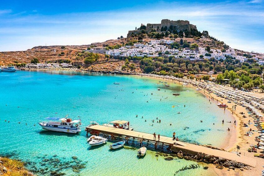 A coastal town with a historic castle on a hill, clear turquoise waters, and a beach with people and boats. 5