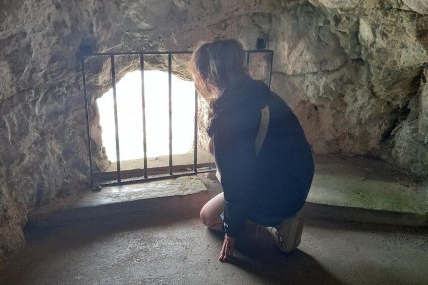 A person crouching inside a stone-walled room with a barred window. 3