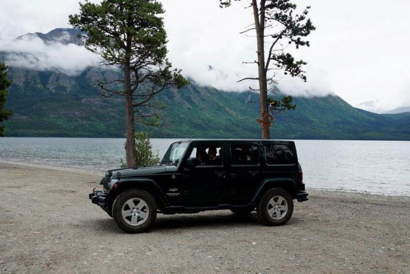 Un véhicule tout-terrain noir est garé sur une plage de galets, avec un lac et des montagnes en arrière-plan. 2