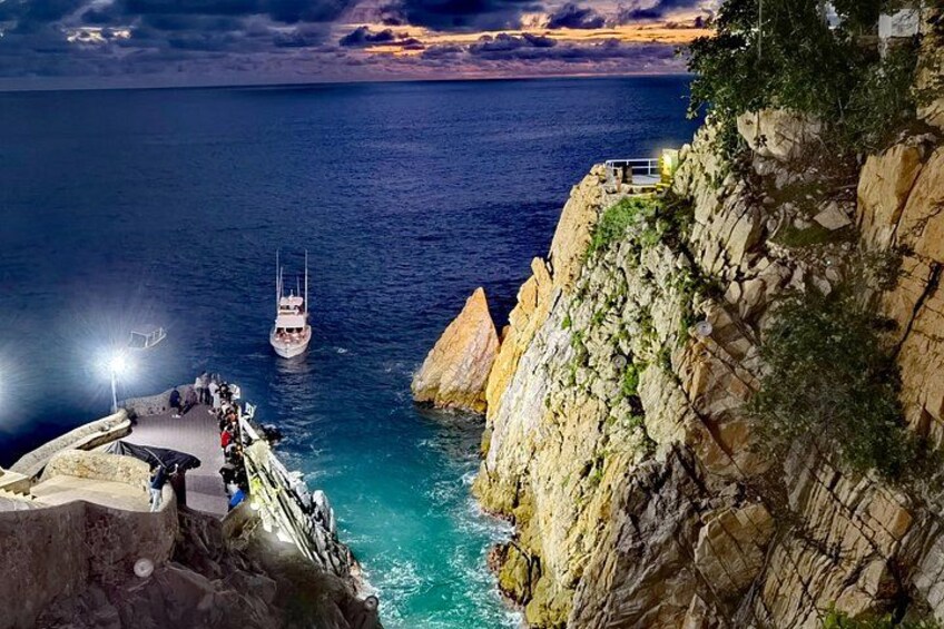 A rocky coastline with a boat anchored near a cliff, a person standing on the edge, and a sunset in the background. 1