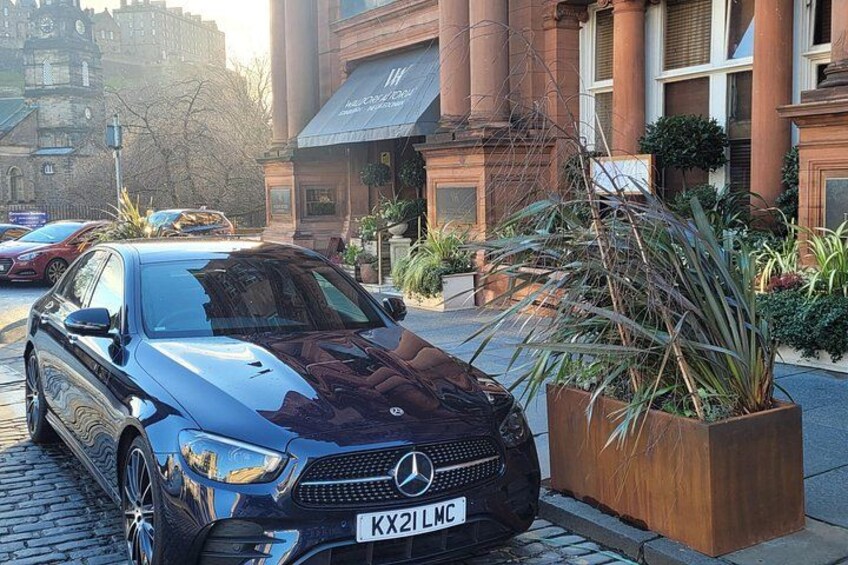Un Mercedes-Benz azul estacionado en una calle adoquinada, con una jardinera grande de plantas altas a su lado. 2