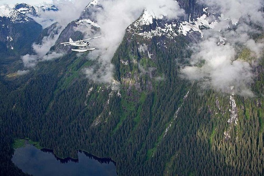 A seaplane flying over a mountainous area with dense forests and a lake. 3