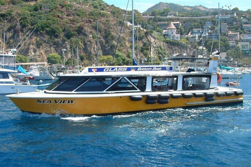 En gul båt med namnet ”SEA VIEW” och texten ”GLASS Bottom Boat” målad på sidan, som seglar på vattnet med en kuststad i bakgrunden. 2