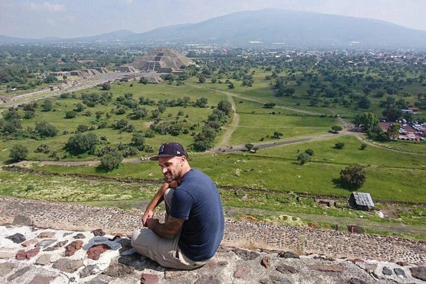 Ein Mann sitzt auf einer Steinmauer und blickt auf eine grüne Landschaft mit alten Ruinen in der Ferne. 2