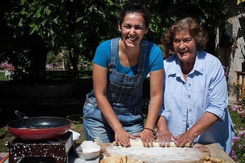 Two people kneeling on the ground, preparing food outdoors with a large cutting board and a red pan. 3