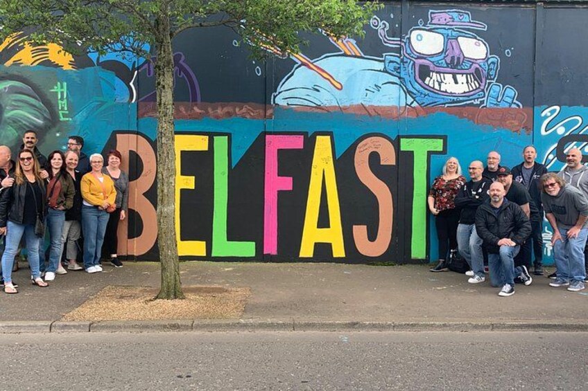 Un gruppo di persone che posa davanti a un murale colorato con la scritta 'BELFAST' in evidenza. 4