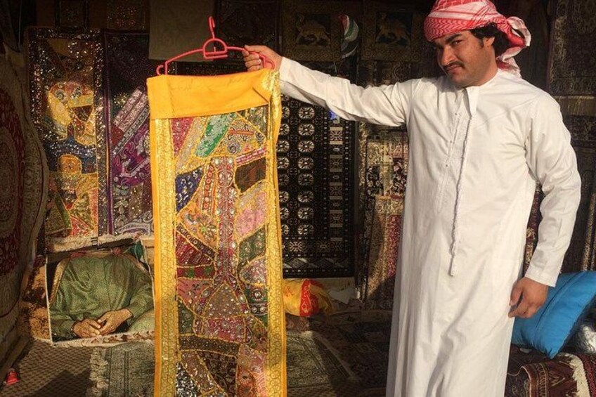 Una persona sostiene un tejido de colores en un perchero, con textiles y alfombras tradicionales de fondo. 5