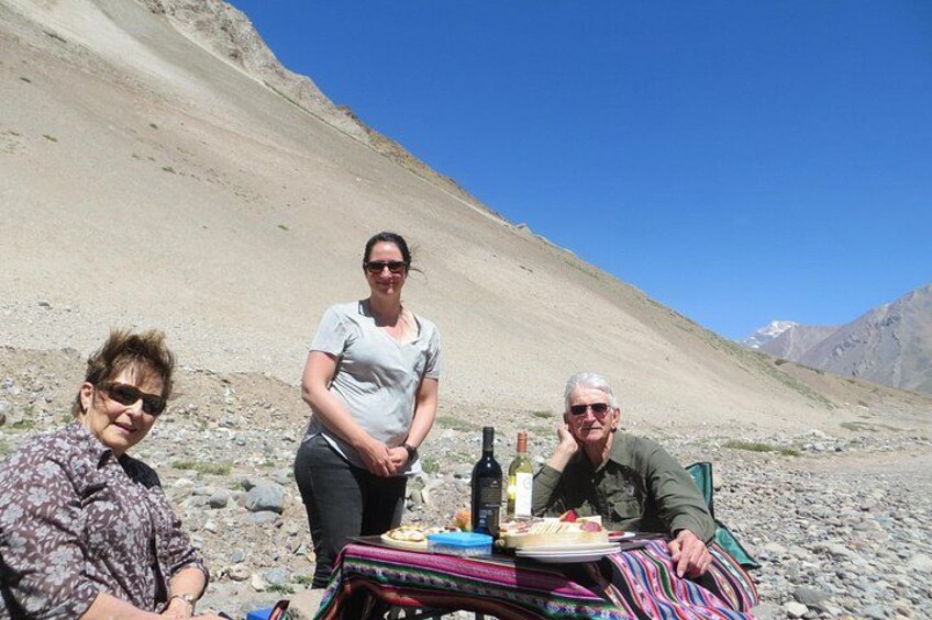 Three people sitting on a blanket with a table in a mountainous area. 5
