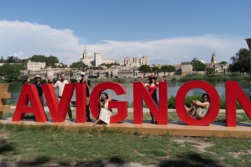 A group of people posing with large red letters spelling 'AVIGNON' in a park with a historic cityscape in the background. 5