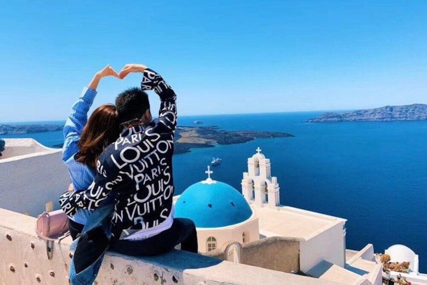 Una pareja posando en un acantilado con vista al mar azul, un edificio blanco y una cúpula azul al fondo. 1