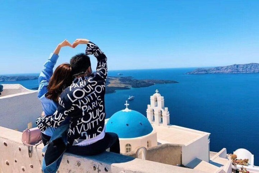 Una coppia che posa formando un cuore, con vista su una chiesa a cupole blu e il mare. 1