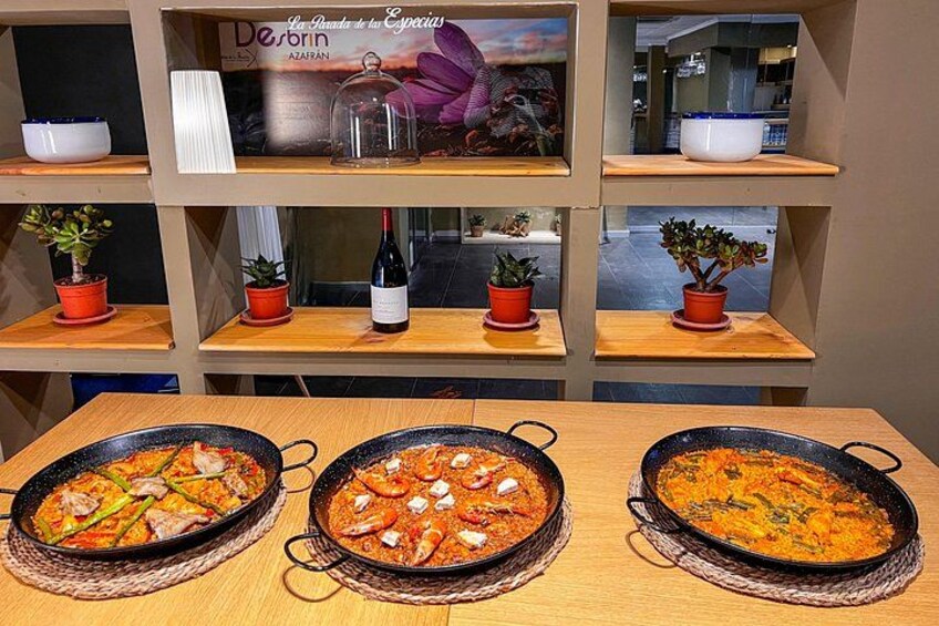 Tres ollas grandes de comida sobre una mesa de madera, rodeadas de estantes con plantas en macetas y un cuadro enmarcado. 3