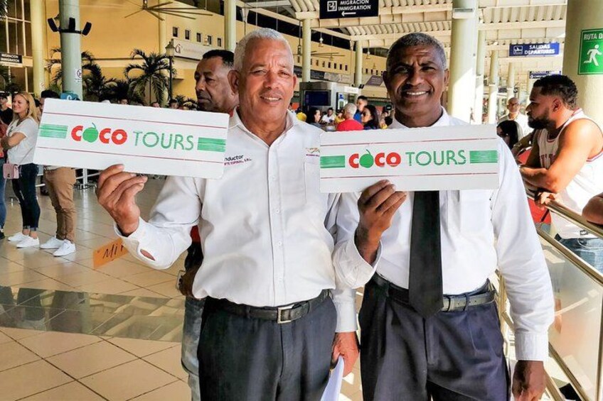 Two individuals holding signs for coco tours in a public indoor space with tiled flooring and other people in the background. 5
