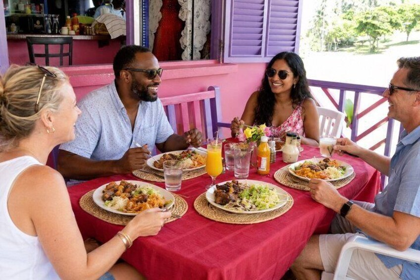 A group of people dining at an outdoor table with a red tablecloth, surrounded by food and drinks. 3