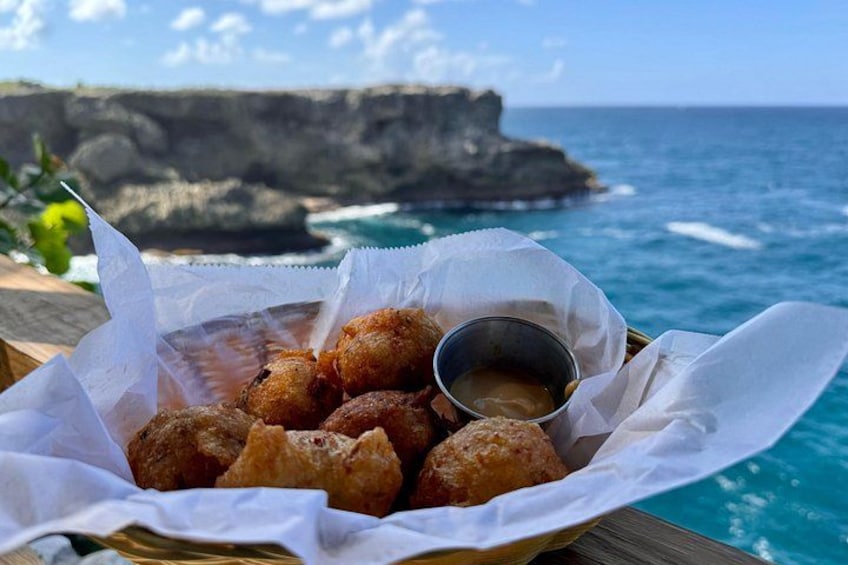 A basket of fried dough balls with dipping sauce, set against a coastal backdrop with cliffs and ocean. 3