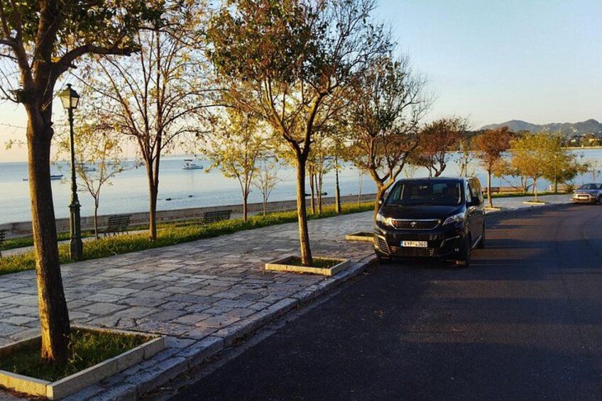 A paved walkway with trees, a parked car, and a body of water in the background. 3
