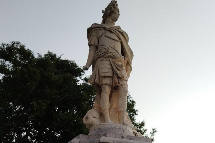 A classical statue of a figure in armor, standing on a pedestal, with trees in the background. 5