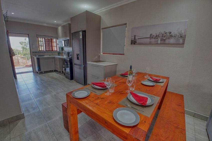 A dining area with a wooden table set for four, a kitchen in the background, and a cityscape painting on the wall. 3