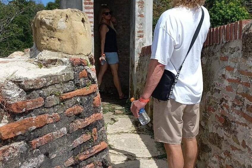 Un hombre de pie sobre un muro de piedra con una estructura de ladrillo al fondo, sosteniendo una botella de agua. 1