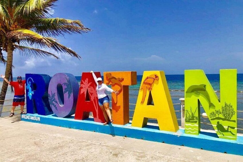 Large colorful letters spelling 'ROATAN' with palm trees and ocean in the background. 2