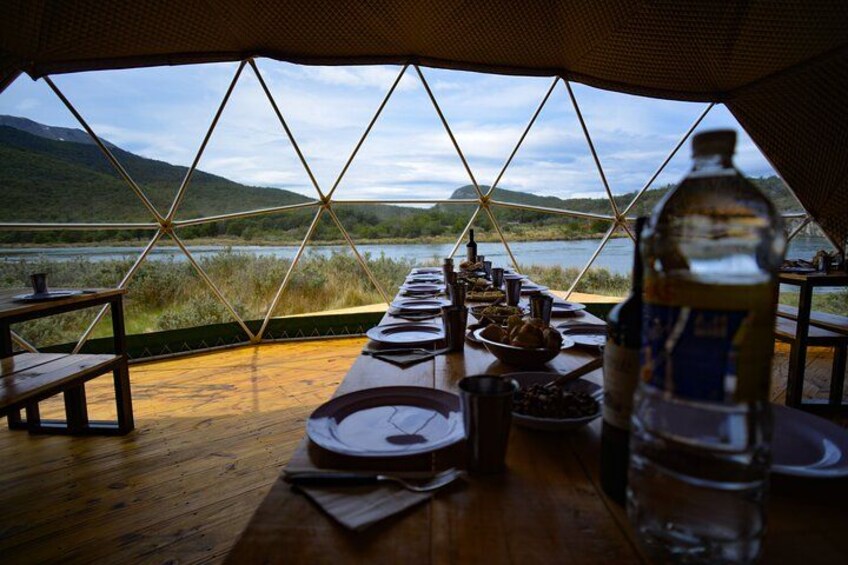 A dining table set up inside a geodesic dome with a view of a lake and mountains. 3