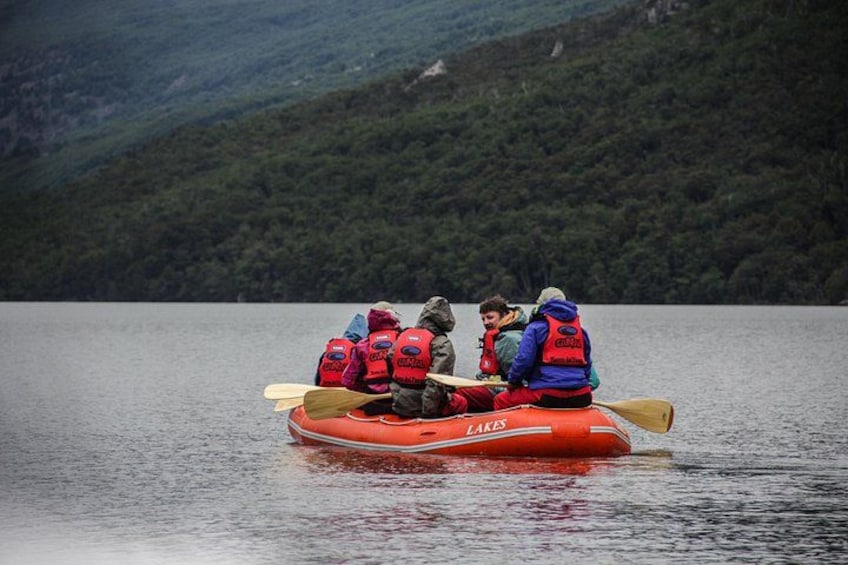 Tres personas en un bote inflable rojo sobre un lago tranquilo, con una montaña boscosa al fondo. 3
