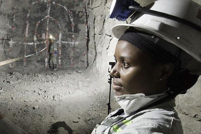 A person in a hard hat and safety vest in an underground setting with visible rock formations and a rope. 1