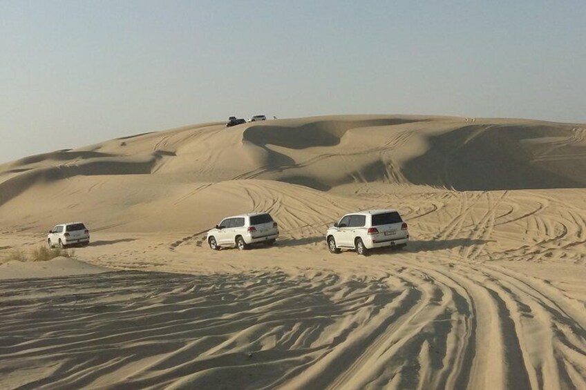 A desert landscape with sand dunes and four white SUVs parked on the sand. 2
