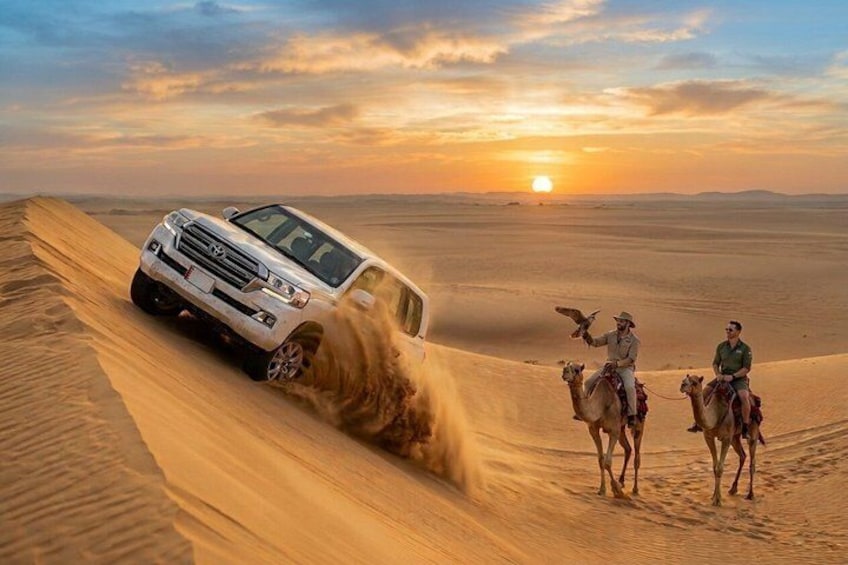 A white SUV driving through a desert with sand dunes, a sunset in the background, and two people riding camels. 1