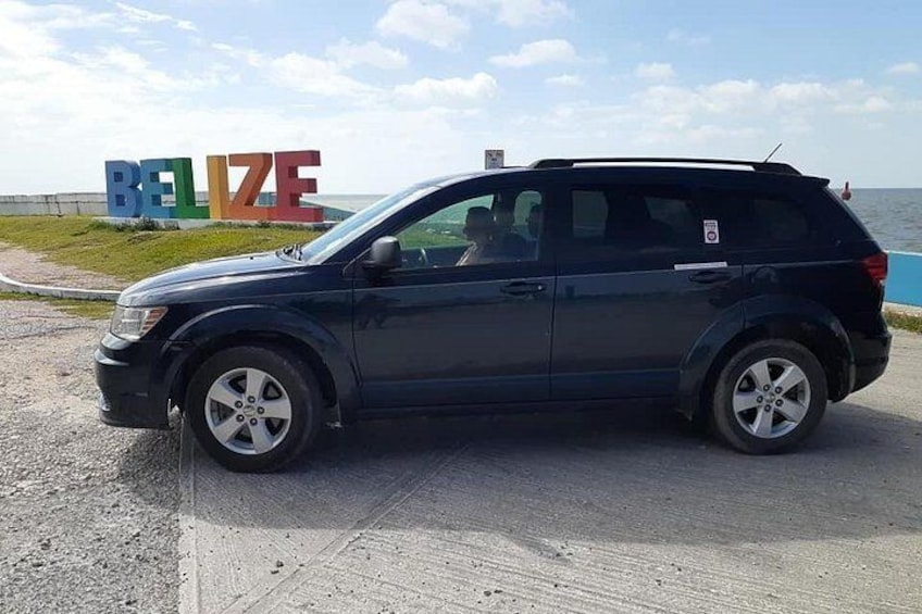 Un SUV de color oscuro estacionado sobre una superficie de concreto cerca de un cuerpo de agua, con un cartel que dice 'BELIZE' al fondo. 3
