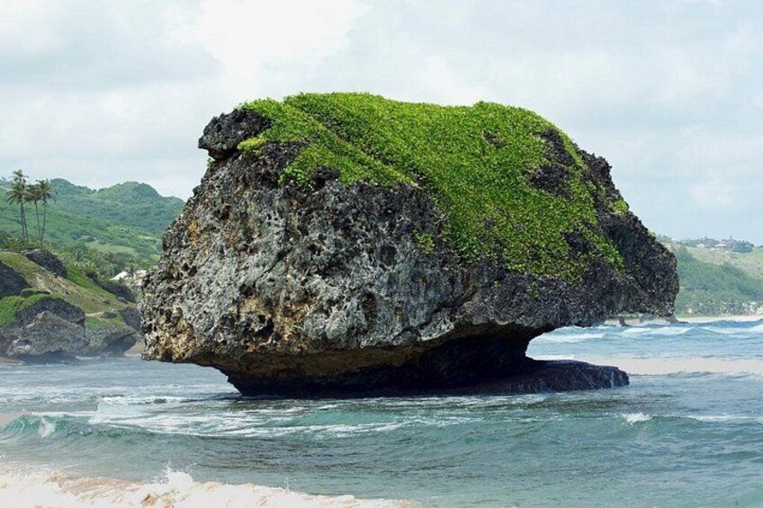 Una gran formación rocosa cubierta de musgo verde, rodeada por olas del océano y una costa distante. 4