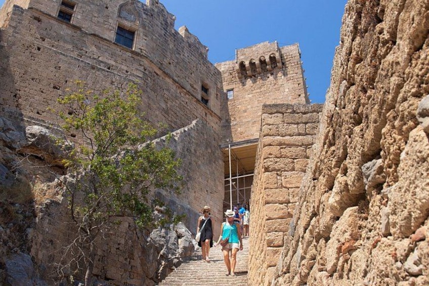 A narrow stone pathway flanked by ancient stone walls, leading to a large fortress-like structure with a tower. 2