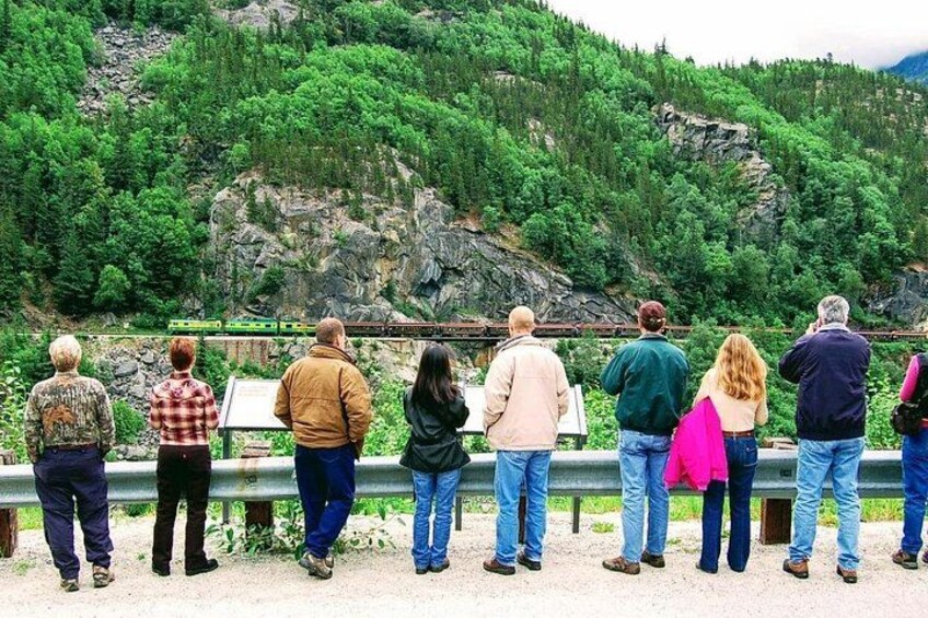 Eine Gruppe von Personen steht an einem Geländer und blickt auf eine gebirgige Landschaft, in der Ferne ist ein Zug zu sehen. 2