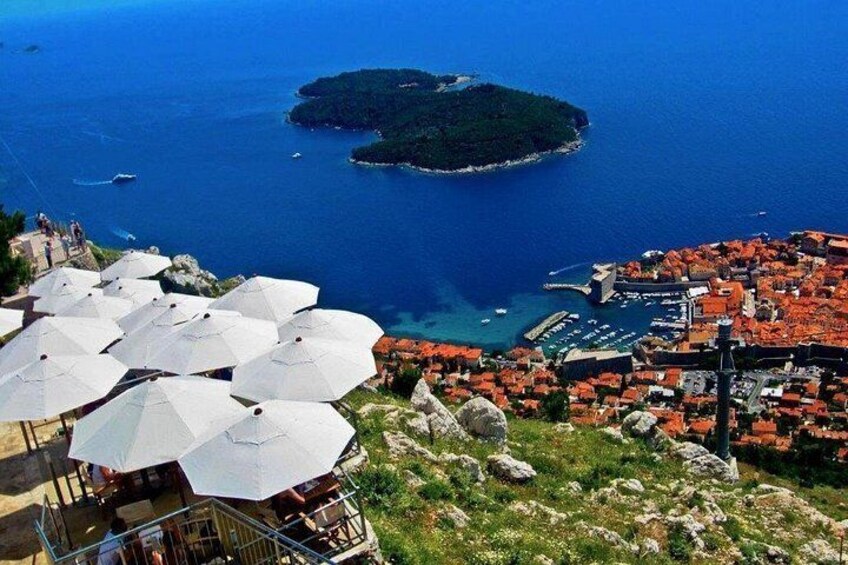 A coastal town with red-roofed buildings, a small island in the sea, and white umbrellas on a hill. 5