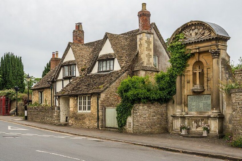 A traditional stone house with a white window trim, a stone wall with greenery, and a small stone structure with a cross. 1