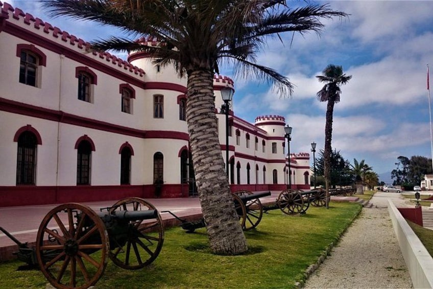 A historic building with red and white walls, palm trees, and old-fashioned carriages. 1