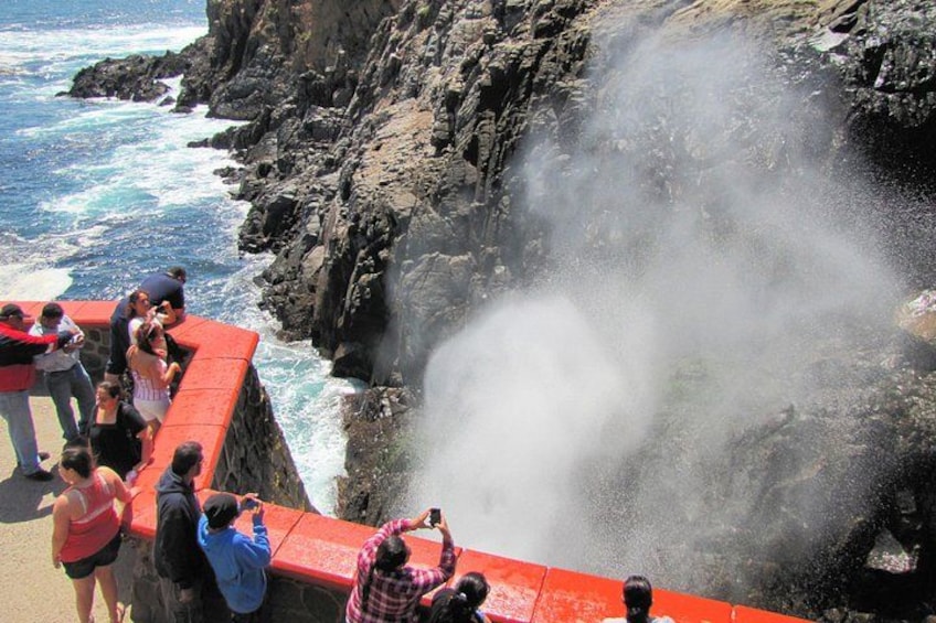 A coastal scene with a rocky cliff, ocean waves, and a group of people observing a geyser. 3