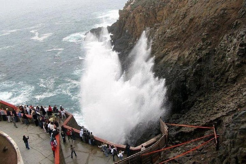 A coastal scene with a large wave crashing against a cliff, a crowd of people observing the event, and a rocky shoreline. 5