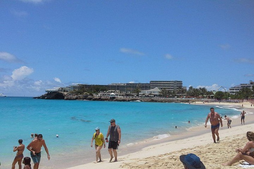 A beach with people walking and swimming, clear turquoise water, and buildings in the background. 5