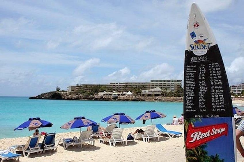 A beach with lounge chairs and umbrellas, a surfboard with a list of arrivals, and a distant building on a small island. 2