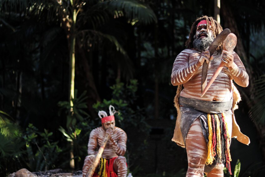 A person in a forested area, wearing traditional attire with a headpiece, holding a wooden object. 4