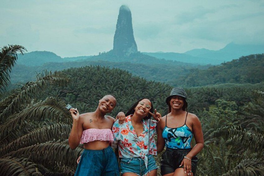 Three people standing in a tropical landscape with dense vegetation and a prominent rock formation in the background. 2