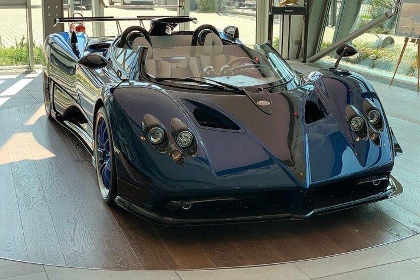 A sleek, dark-colored sports car with a low profile and distinctive front design, parked indoors. 1