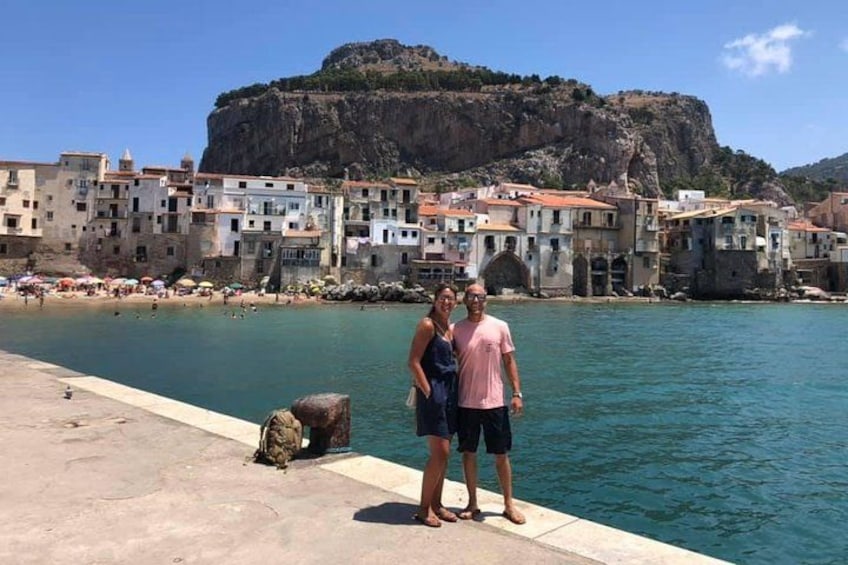 A coastal town with buildings, a rocky cliff, and a calm sea. 1