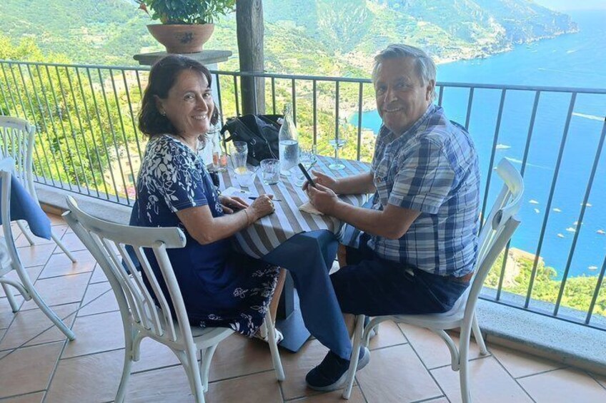 Two people seated at a table on a balcony overlooking a body of water and greenery. 3