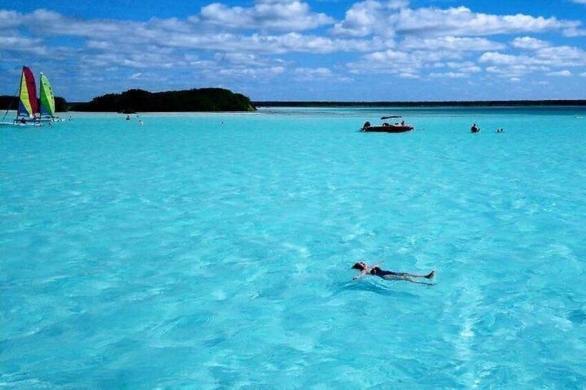 A person swimming in clear turquoise water, a sailboat, and a small island in the background. 1