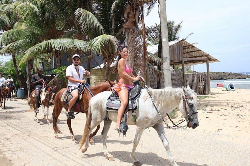 People riding horses on a beach path with palm trees and a wooden structure in the background. 5